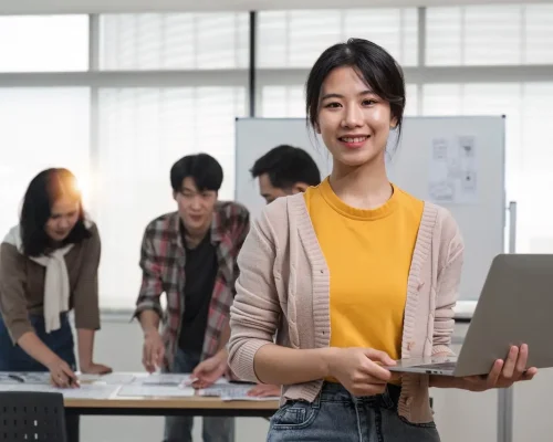 young Asian female marketing assistant is working on her work on her laptop at her desk while her coworkers are working in the room